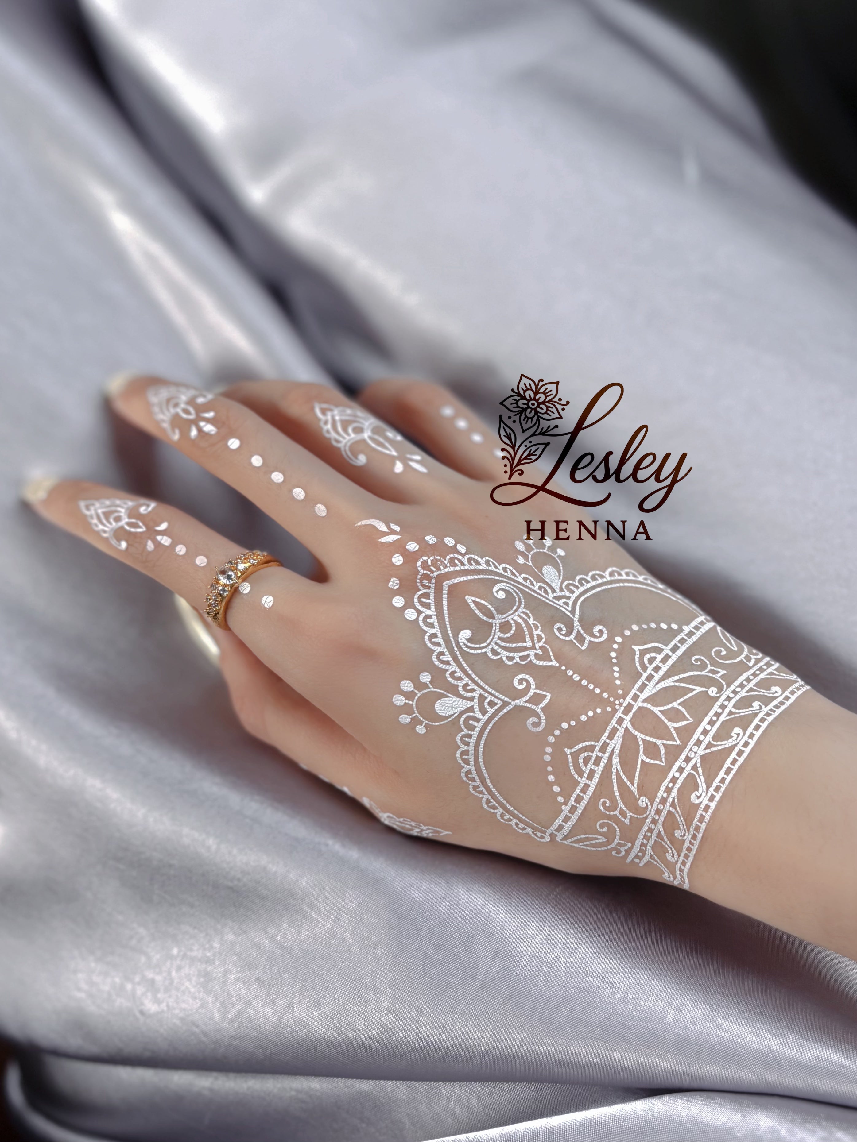 Hand with white henna design on a gray fabric background, featuring the 'Lesley Henna' brand. Bridal white henna sticker inspired by crown motifs