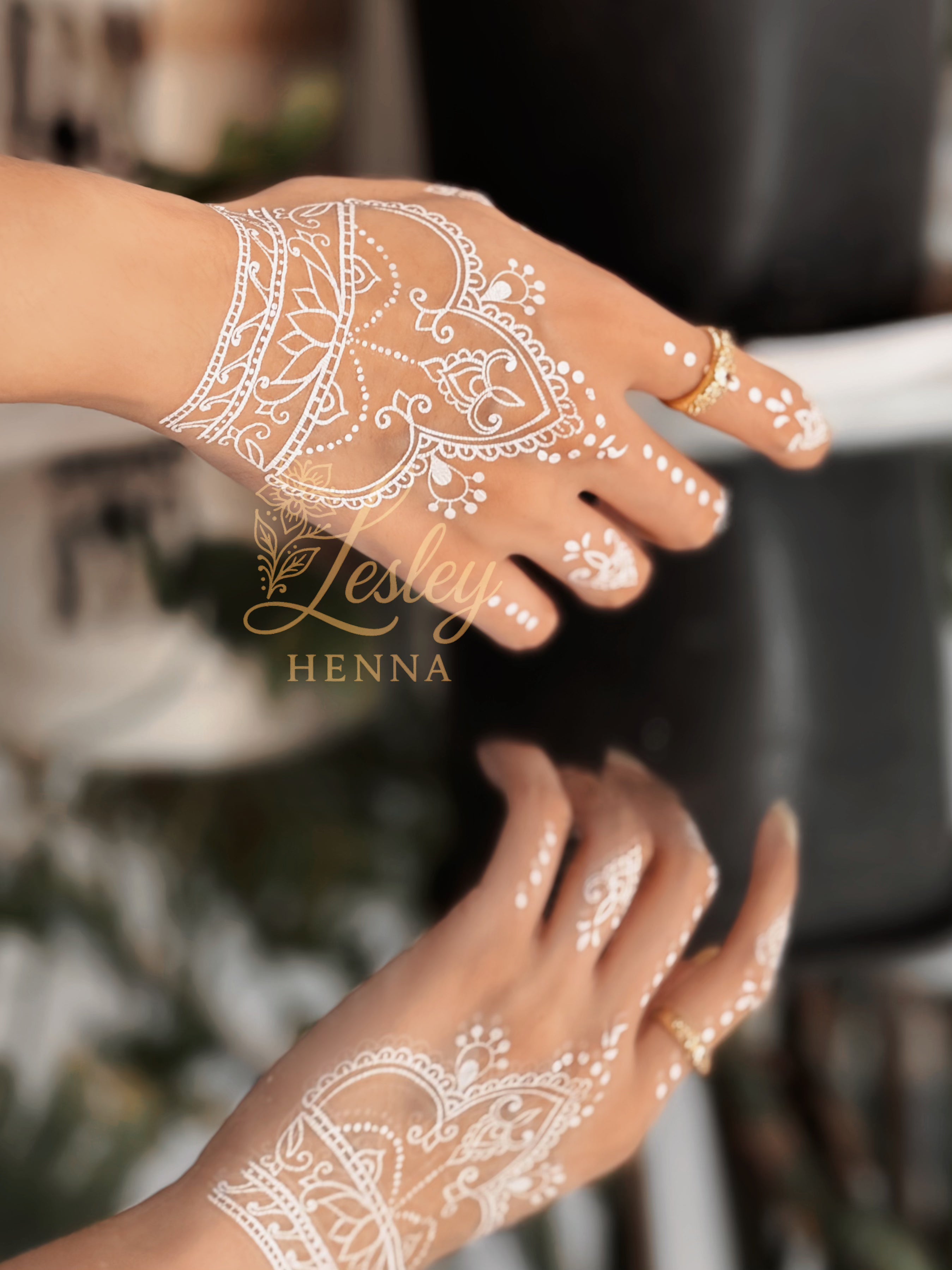 Hands with white henna designs on a blurred background with 'Lesley Henna' branding.