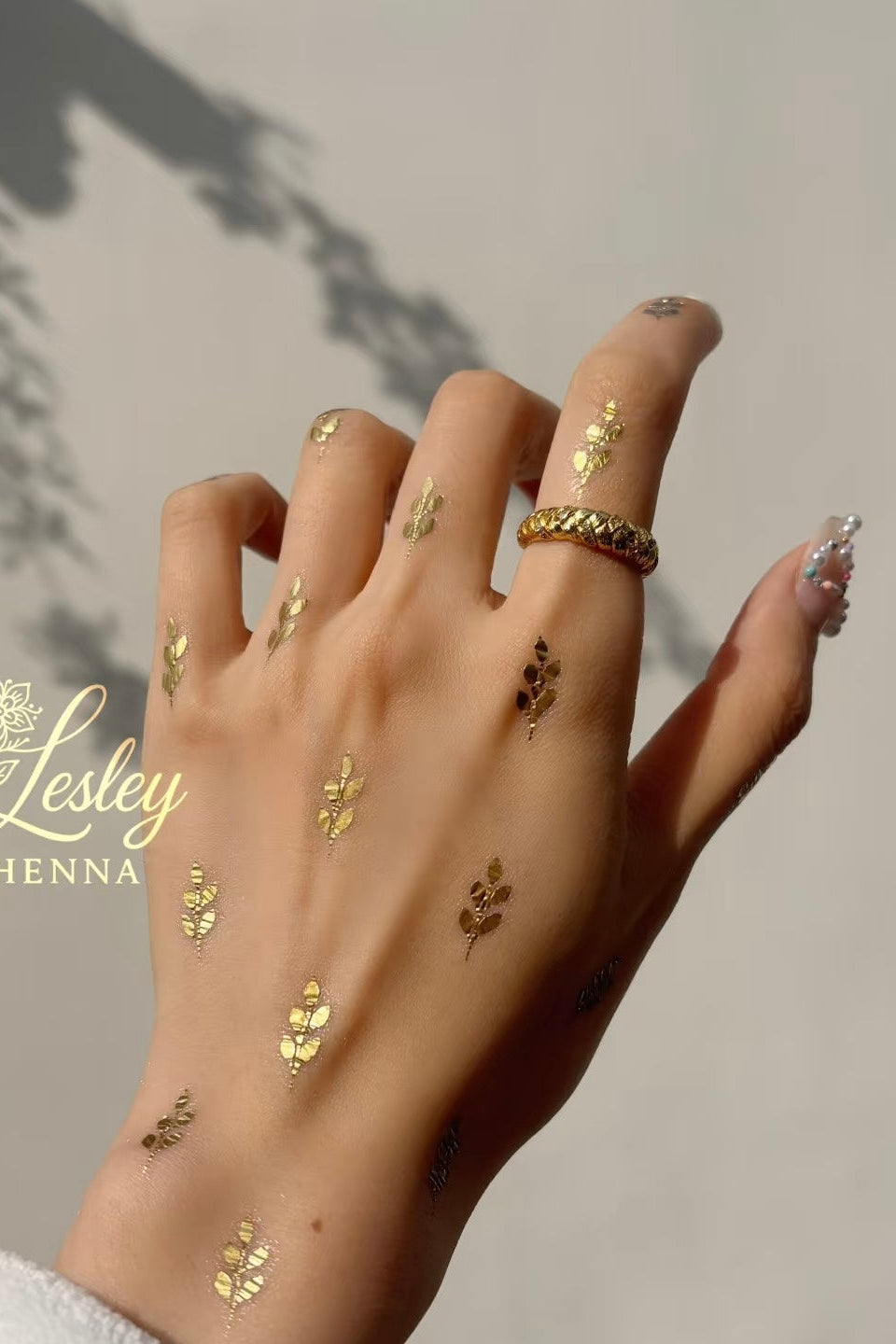 Hand with gold henna designs on a neutral background, featuring the Lesley Henna logo.